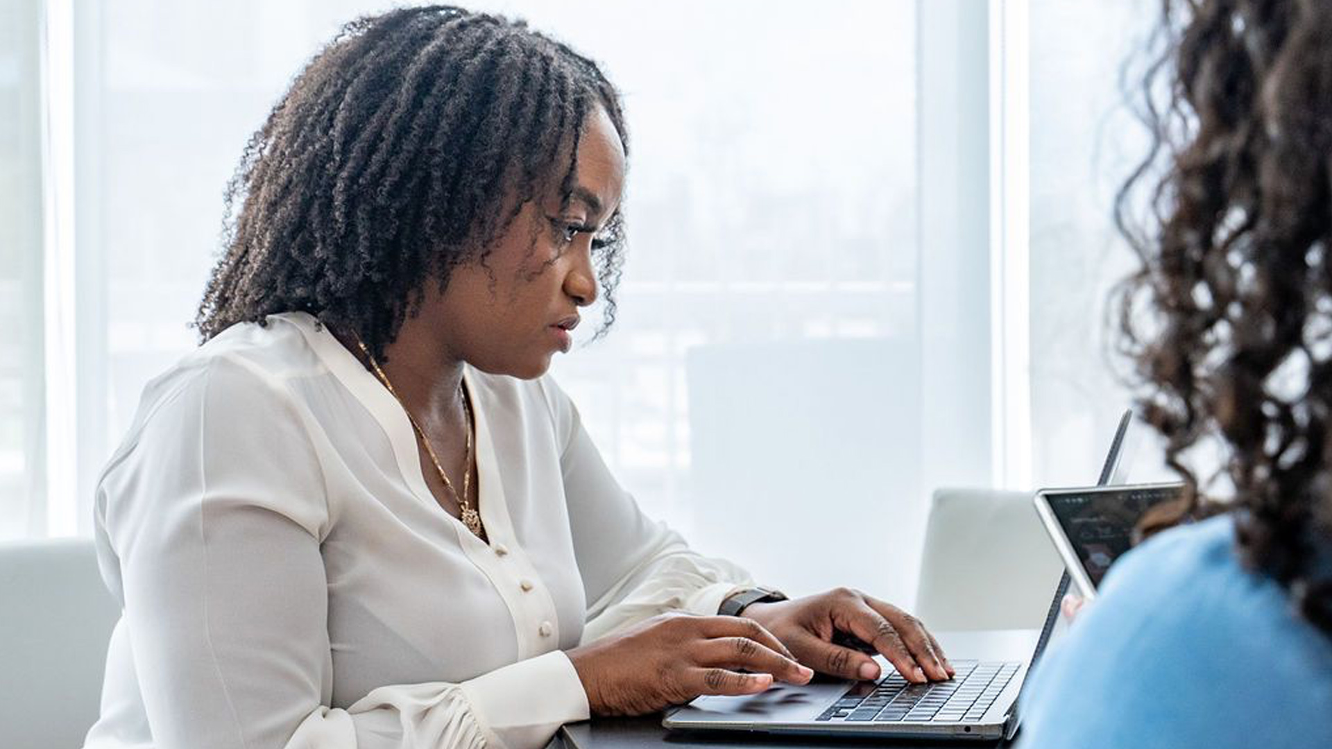 Woman in a white blouse typing on a laptop at a sunlit office table, with a colleague holding a tablet nearby.
